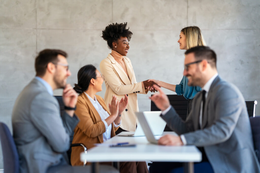 Two business professionals standing and shaking hands while their workers applaud them.